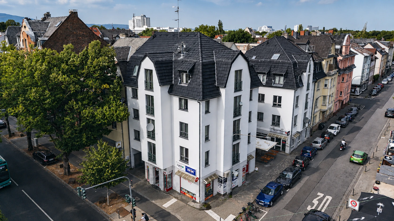 Luftaufnahme eines weißen Eckhauses mit dunklem Ziegeldach und Gauben an einer Straßenkreuzung; daneben Bäume, parkende Autos und die Nachbarbebauung.