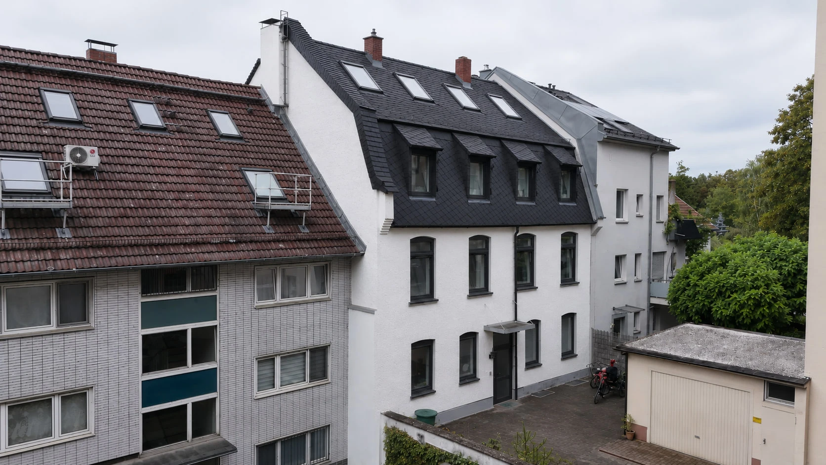 BACKYARD 24 von der Hofseite, schräg von oben: die weiße Fassade mit dunklem Schieferdach und Gauben, rechts daneben ein flaches Garagengebäude und ein gepflasterter Hof.