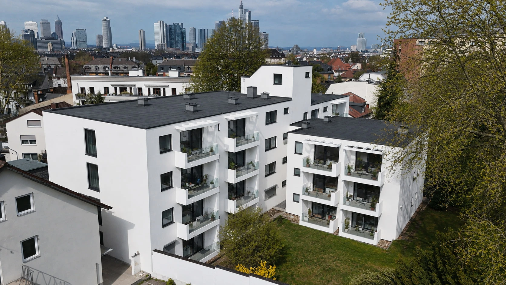 Das Projekt SOUTH25 aus der Luft von oben: zwei weiße Baukörper mit dunklen Dächern und Balkonen, dazwischen eine Rasenfläche; im Hintergrund die Dächer des Viertels und die Frankfurter Skyline.