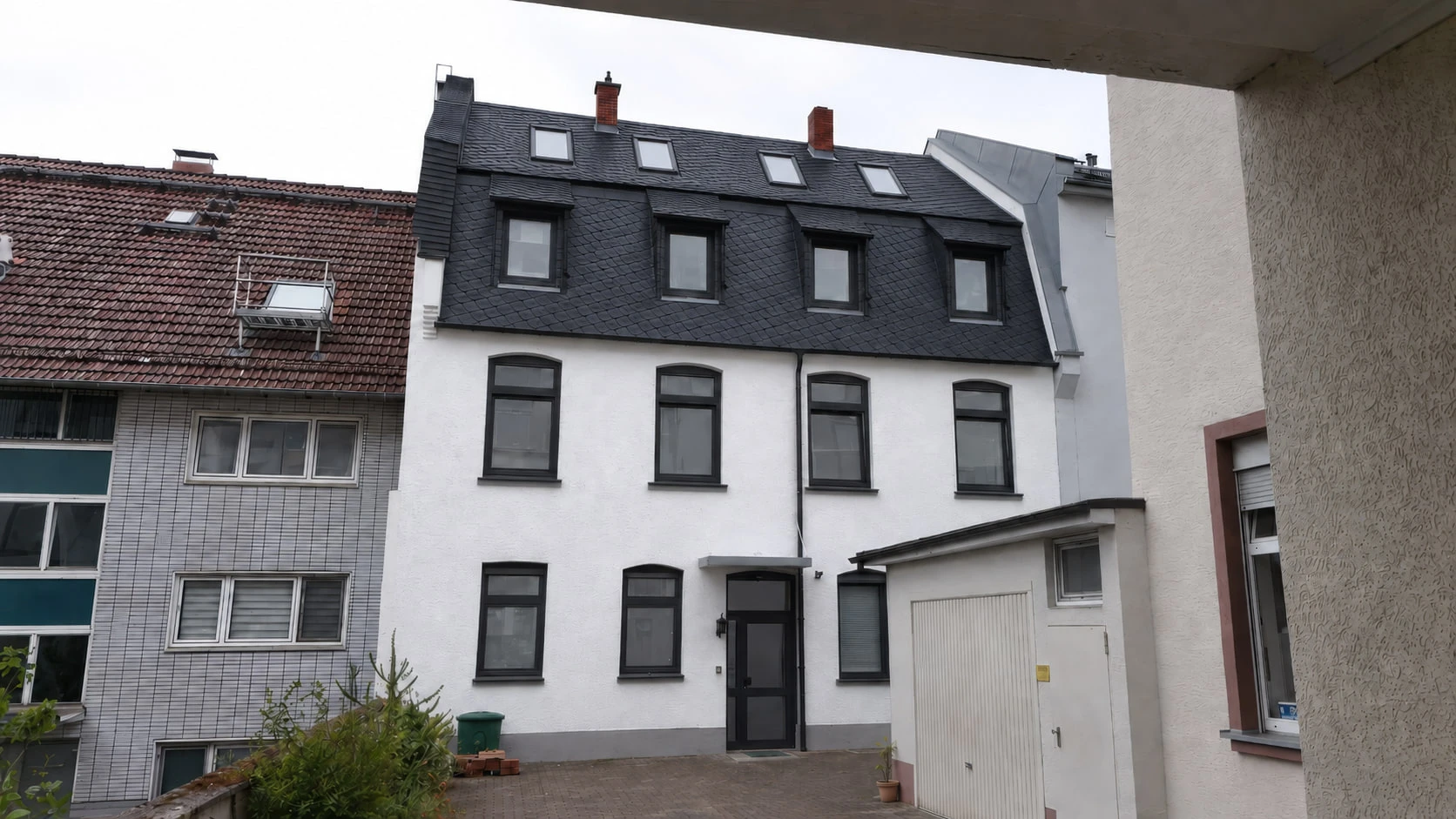 BACKYARD 24 frontal von der Hofseite: weiße Putzfassade mit dunklem Schieferdach, hohen Fenstern in dunklen Rahmen und dem dunklen Hauseingang; rechts die weiße Garage, links das Nachbarhaus.