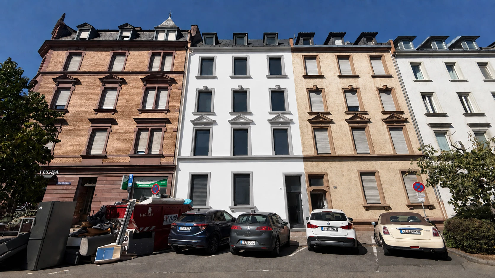 Das Projekt N°36 von der Straße aus: die weiße Putzfassade mit gleichmäßigen Fensterreihen zwischen dem roten Klinkerbau links und den hellen Nachbarhäusern rechts, davor parkende Autos.