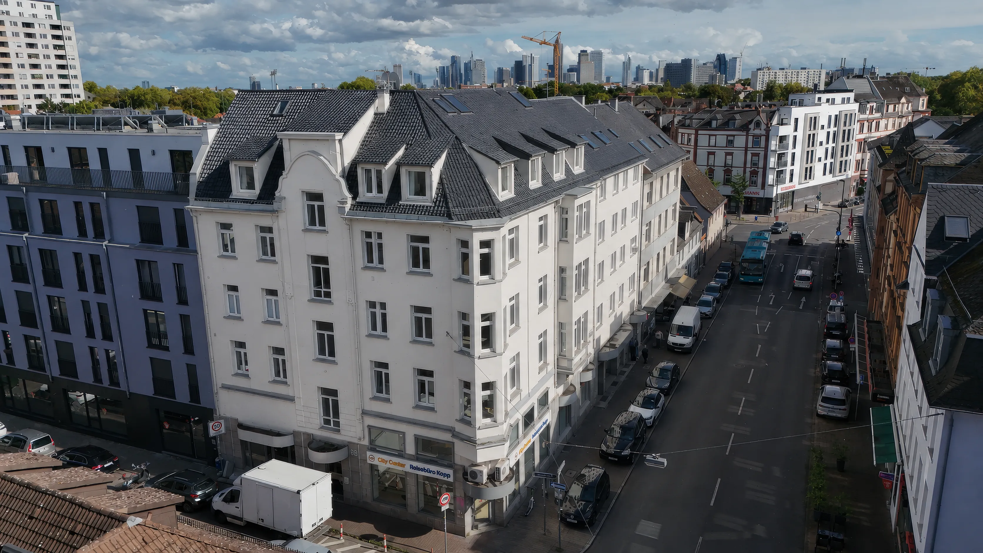 Luftaufnahme des Eckhauses CORNER 36: ein hell verputzter Altbau mit abgeschrägter Gebäudeecke, dunklem Schieferdach, Gauben und Dachflächenfenstern; im Erdgeschoss Ladenlokale, entlang der Straße parkende Autos, im Hintergrund Baumreihen und die Frankfurter Skyline.
