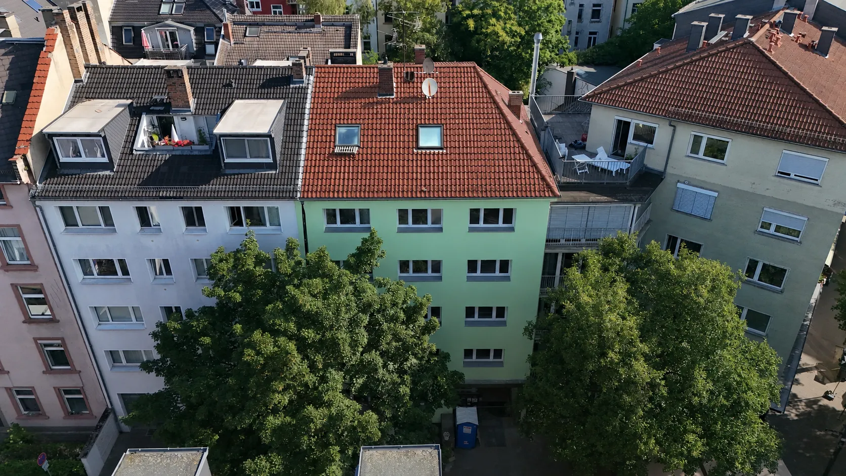 Luftaufnahme des Projekts GREEN FOUR: ein mintgrün verputztes Mehrfamilienhaus mit rotem Ziegel-Walmdach und zwei Dachfenstern, eingebaut in die Häuserzeile; davor zwei große Laubbäume, ringsum die Dächer des Viertels.