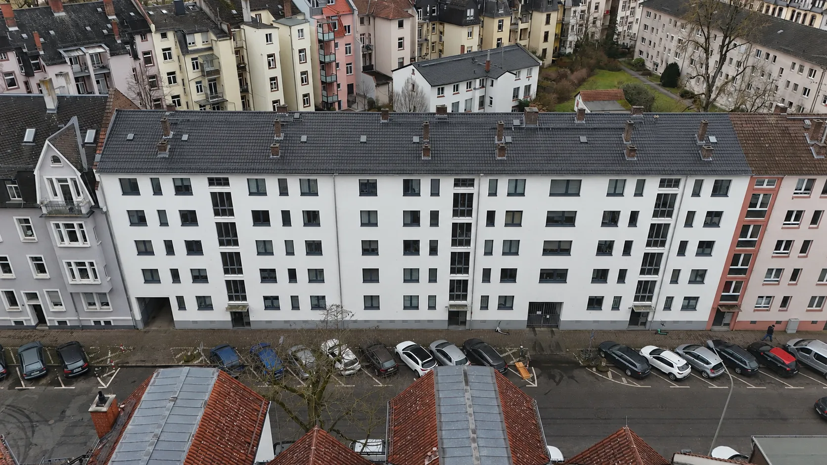 Luftaufnahme des Projekts ENSEMBLE 27: eine lange weiße Wohnzeile mit dunklem Dach entlang der Straße, davor parkende Autos; dahinter die Dächer des Viertels und Grünflächen.
