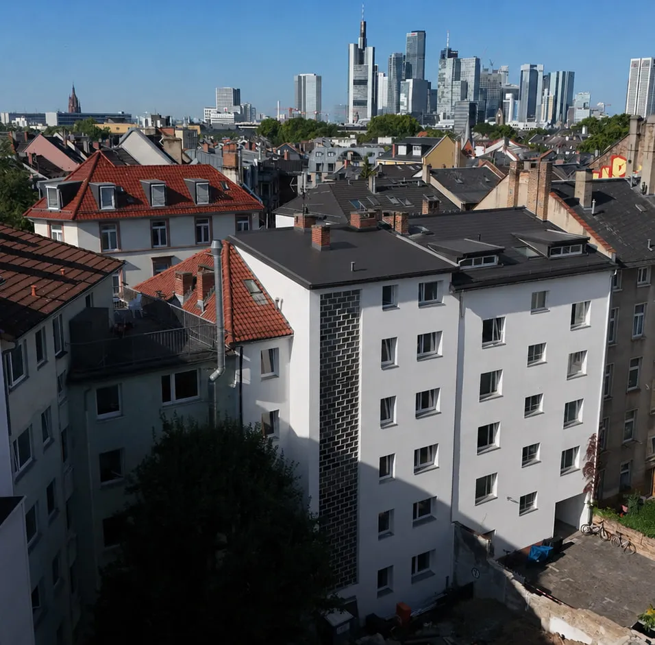 Das Projekt GREEN FOUR von der Hofseite: heller Putzbau mit dunkler Klinkerkante an der Ecke, dahinter die Dächer des Viertels und die Frankfurter Skyline.