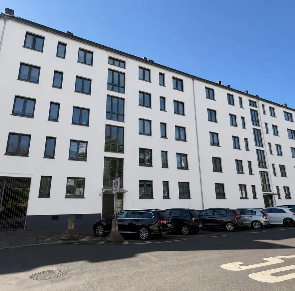 Das Projekt ENSEMBLE 27 von der Straße aus: die lange weiße Wohnzeile mit gleichmäßigen Fensterreihen, davor die Fahrbahn mit der aufgemalten Tempo-30-Markierung und parkenden Autos.